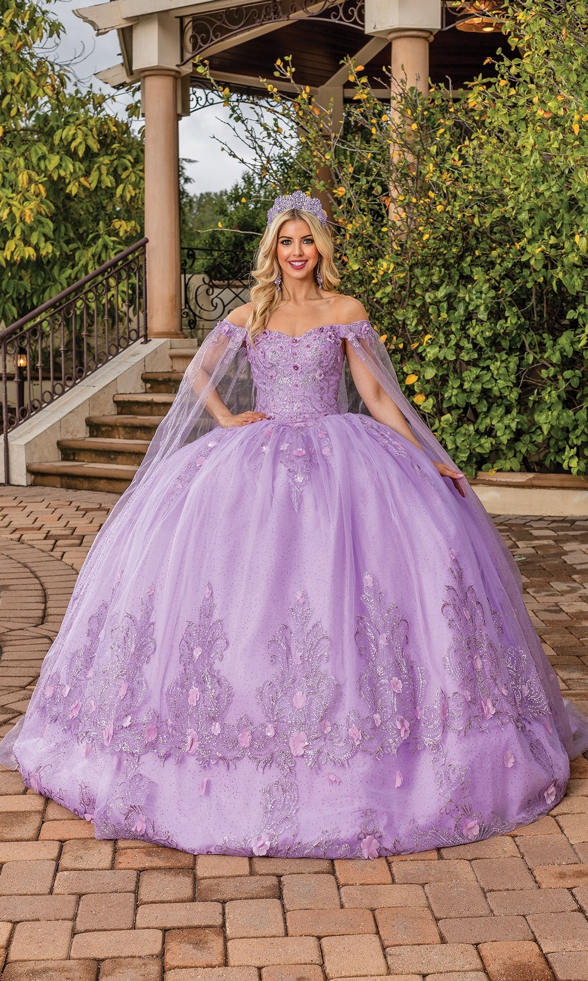 A woman in the Quince Dress with Cape Sleeves: Dancing Queen 1676, featuring lavender fabric and floral embellishments, stands outside on a brick path before greenery and stairs, wearing a tiara.